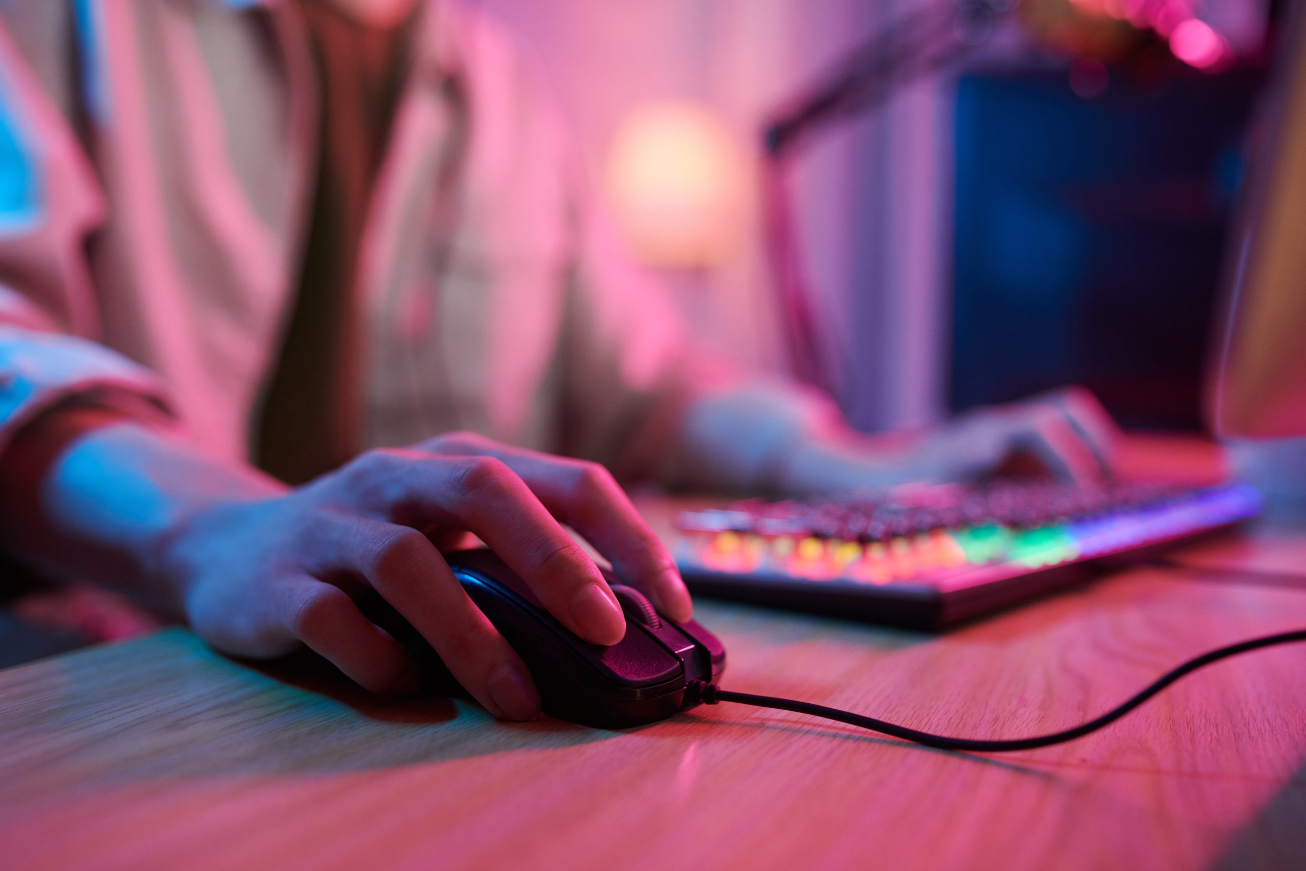 Pessoa segurando um mouse, com uma iluminação rosa e um teclado ao fundo, simulando o uso do computador.
